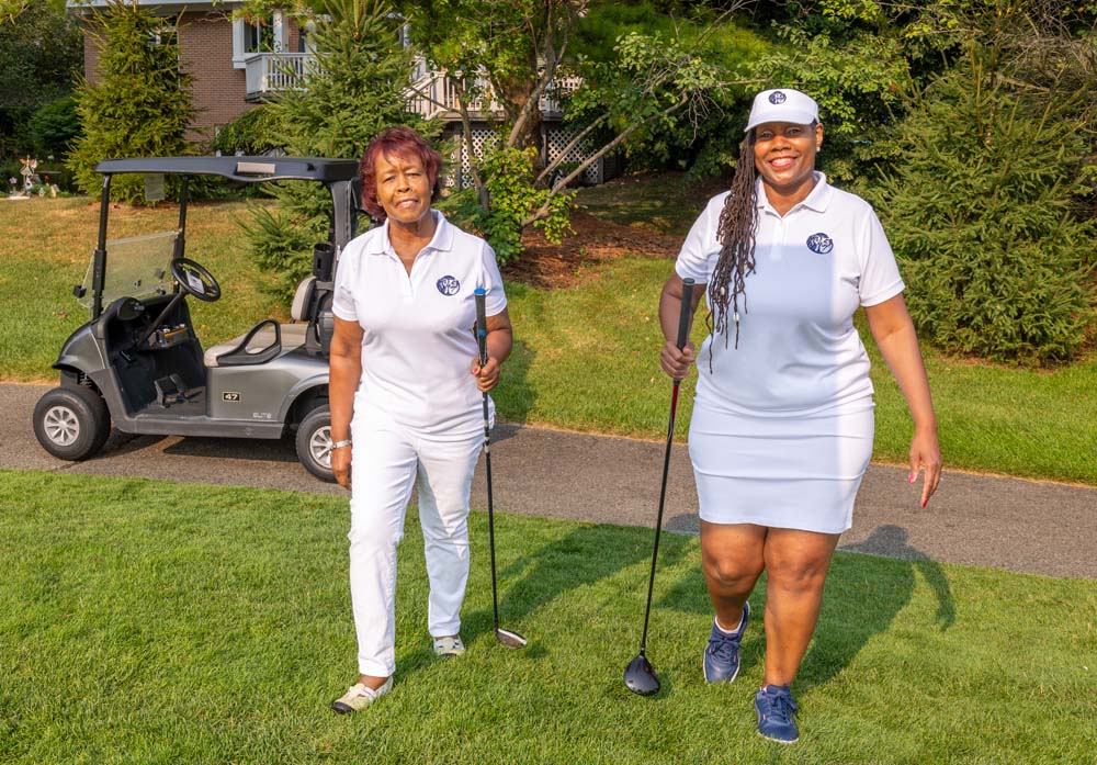 Two women in golf attire standing on a golf course with a golf cart in the background.